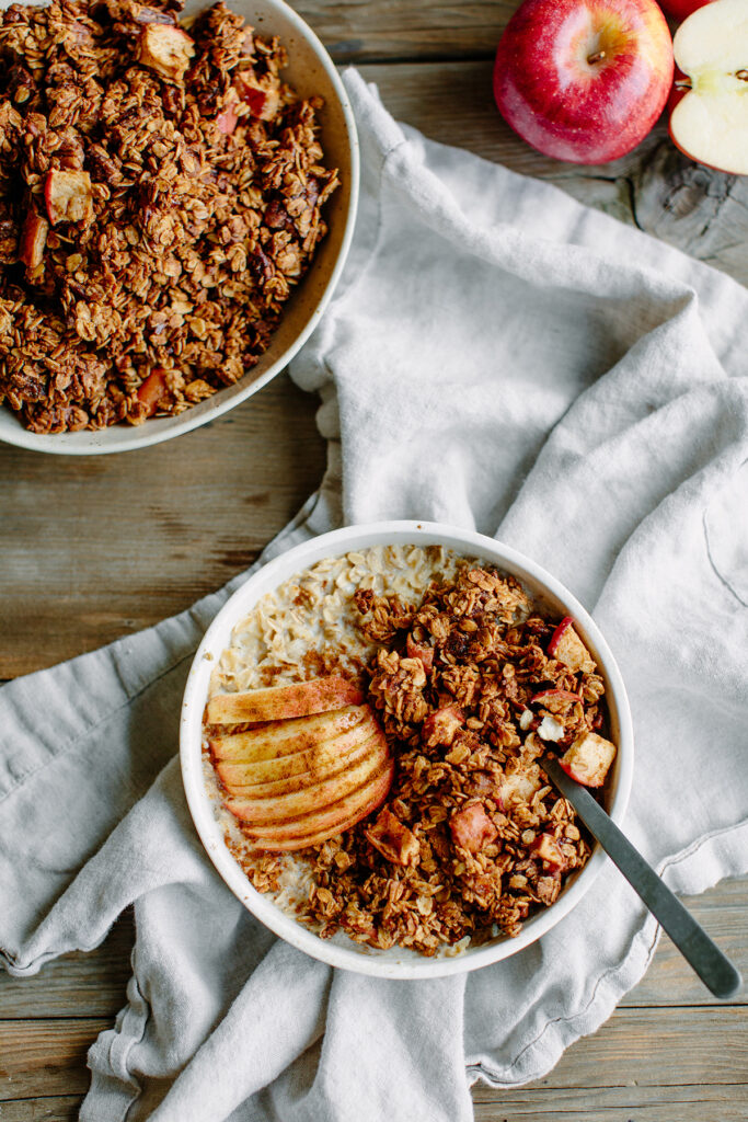 A bowl of oatmeal topped with baked apple slices and granola sits on a light cloth next to a larger bowl of granola and a red apple on a wooden table. A spoon rests in the oatmeal bowl.