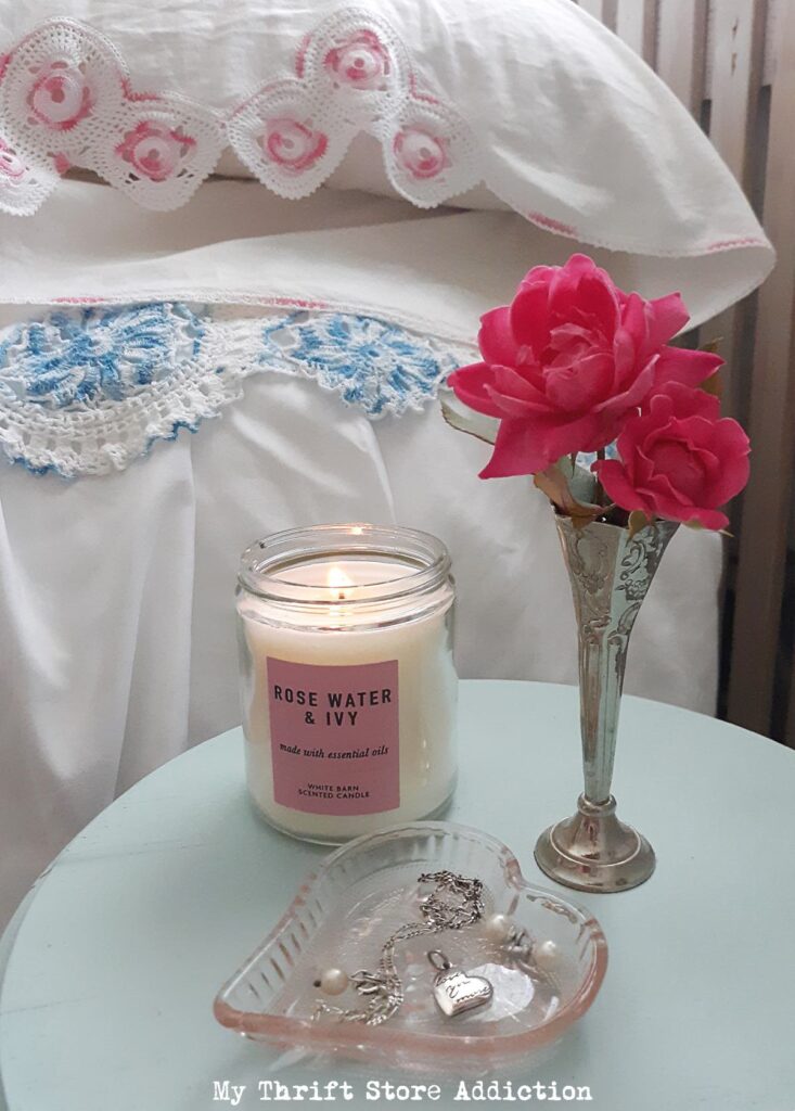 Closeup of two vintage pillow cases with crocheted trim behind a lit scented candle and a vintage silver bud vase with two roses