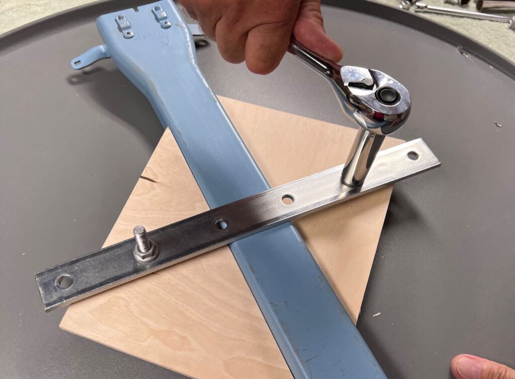 closeup of underside of the swing seat showing a mending plate across the satellite arm with bolts coming through it. a hand is using a ratchet to tighten the nut on the bolt. with a triangle of wood on each side of the arm which holds the arm in place side to side.