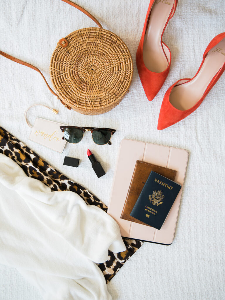 Flat lay of travel essentials on a white surface: a round wicker bag, red high heels, sunglasses, lipstick, an animal-print scarf, a notebook, a U.S. passport, and a white iPad case.