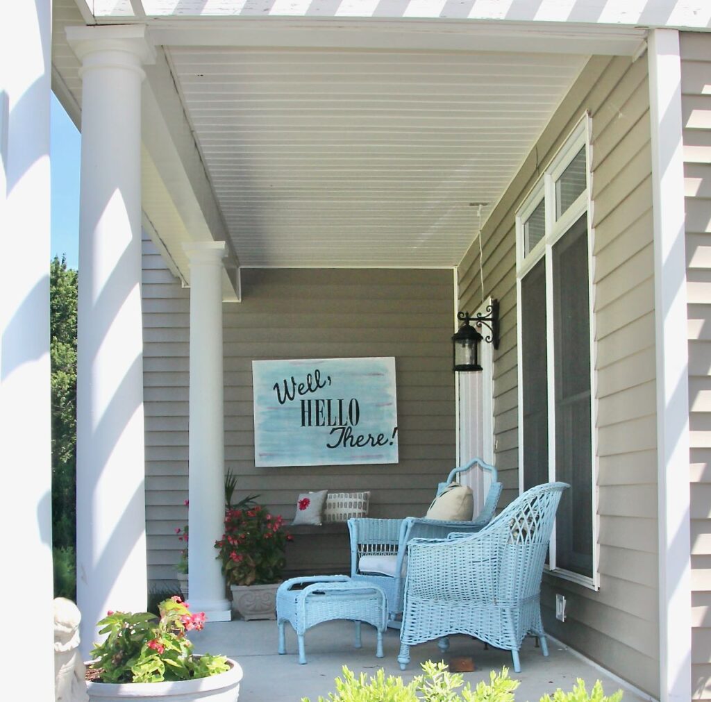 view past wicker furniture on one side and round columns on the other down the length of the front porch of a very large stretched canvas sign that reads "Well, Hello there!" on a black wall of siding adjacent to the front door