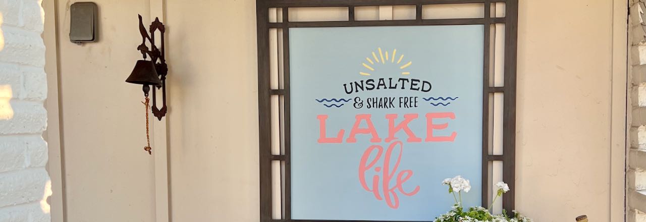 wide closeup of the finished patio sign "unsalted & Shark Free Lake Life" hanging outside with a dinner bell next to it and a planted pot. in front of one corner