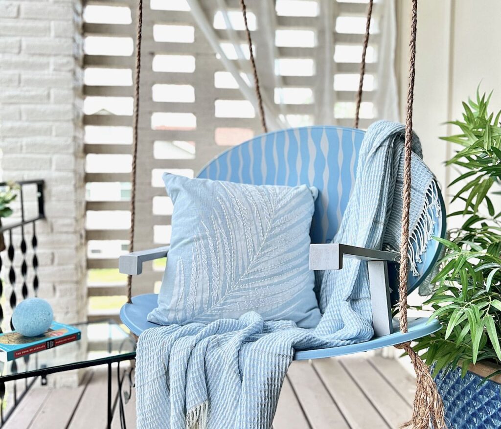 Finished Repurposed Satellite Dish Swing hanging on a balcony next to a green plant with a light throw and accent pillow in the swing