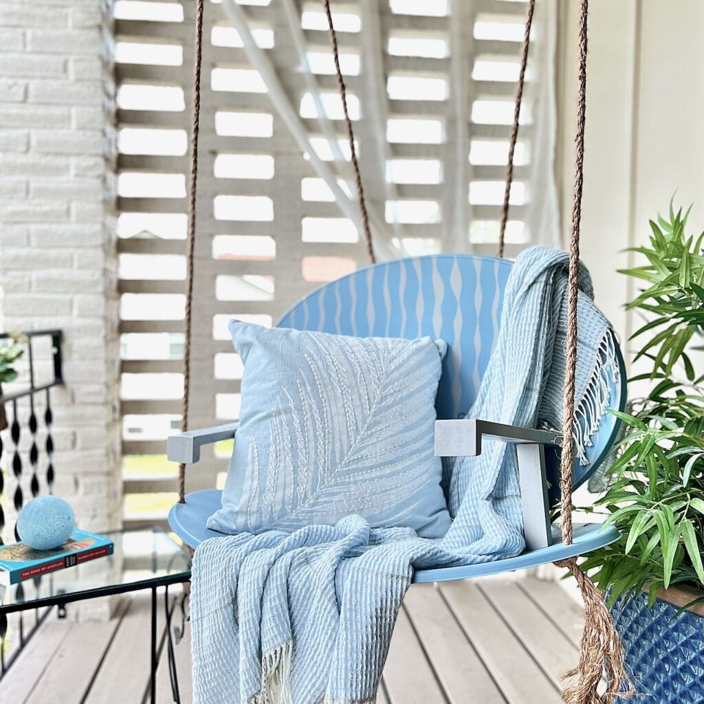 Satellite swing shown hanging in front of a brick screen with sheer curtain tied back. The swing has a light blue throw and a light blue pillow