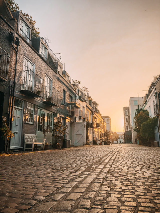 A quiet cobblestone street lined with charming townhouses, potted plants, and balconies, bathed in warm golden light during sunset. The scene feels peaceful and inviting.