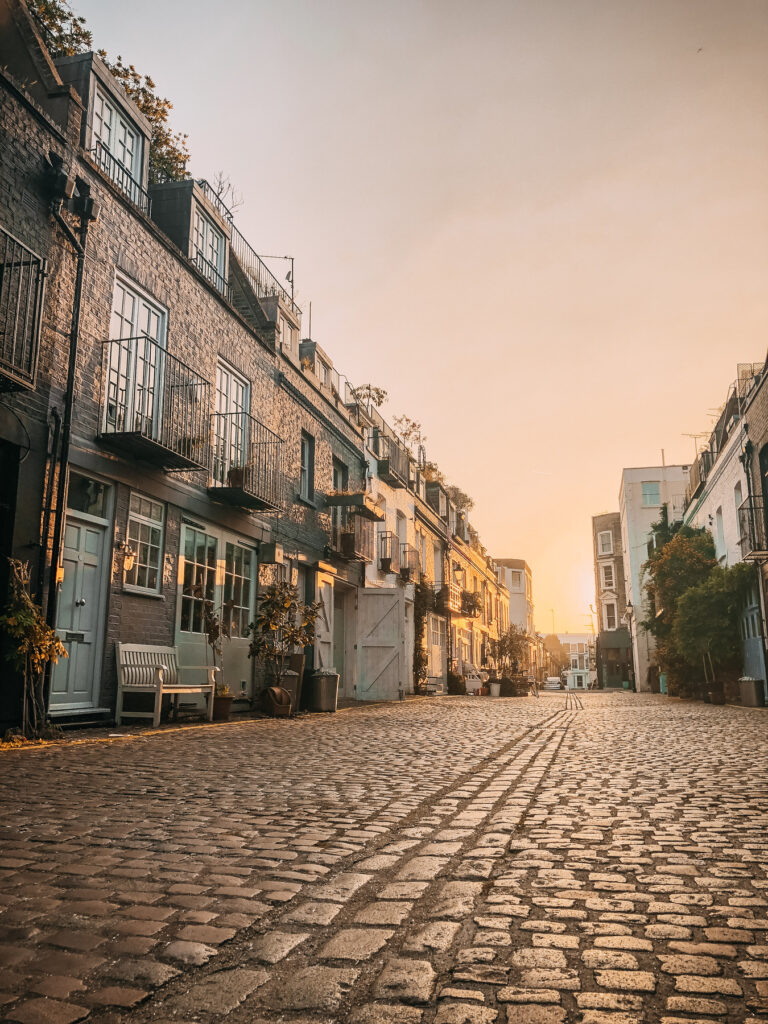 A cobblestone street lined with brick buildings and balconies, bathed in warm golden sunlight at sunset. The street is empty, and the sky glows softly, creating a peaceful, serene atmosphere.