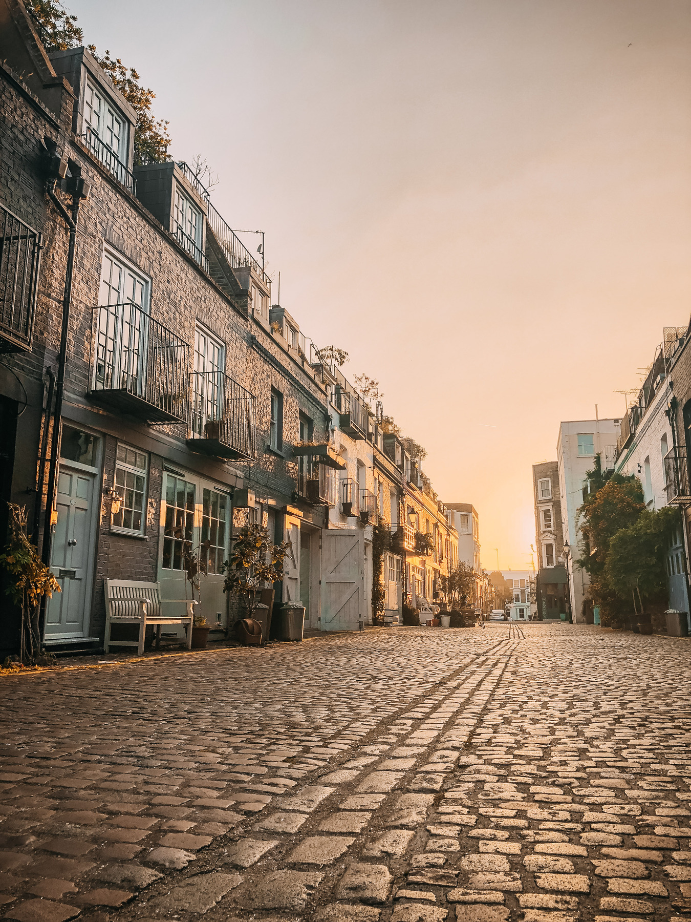 A cobblestone street lined with brick buildings and balconies, bathed in warm golden sunlight at sunset. The street is empty, and the sky glows softly, creating a peaceful, serene atmosphere.