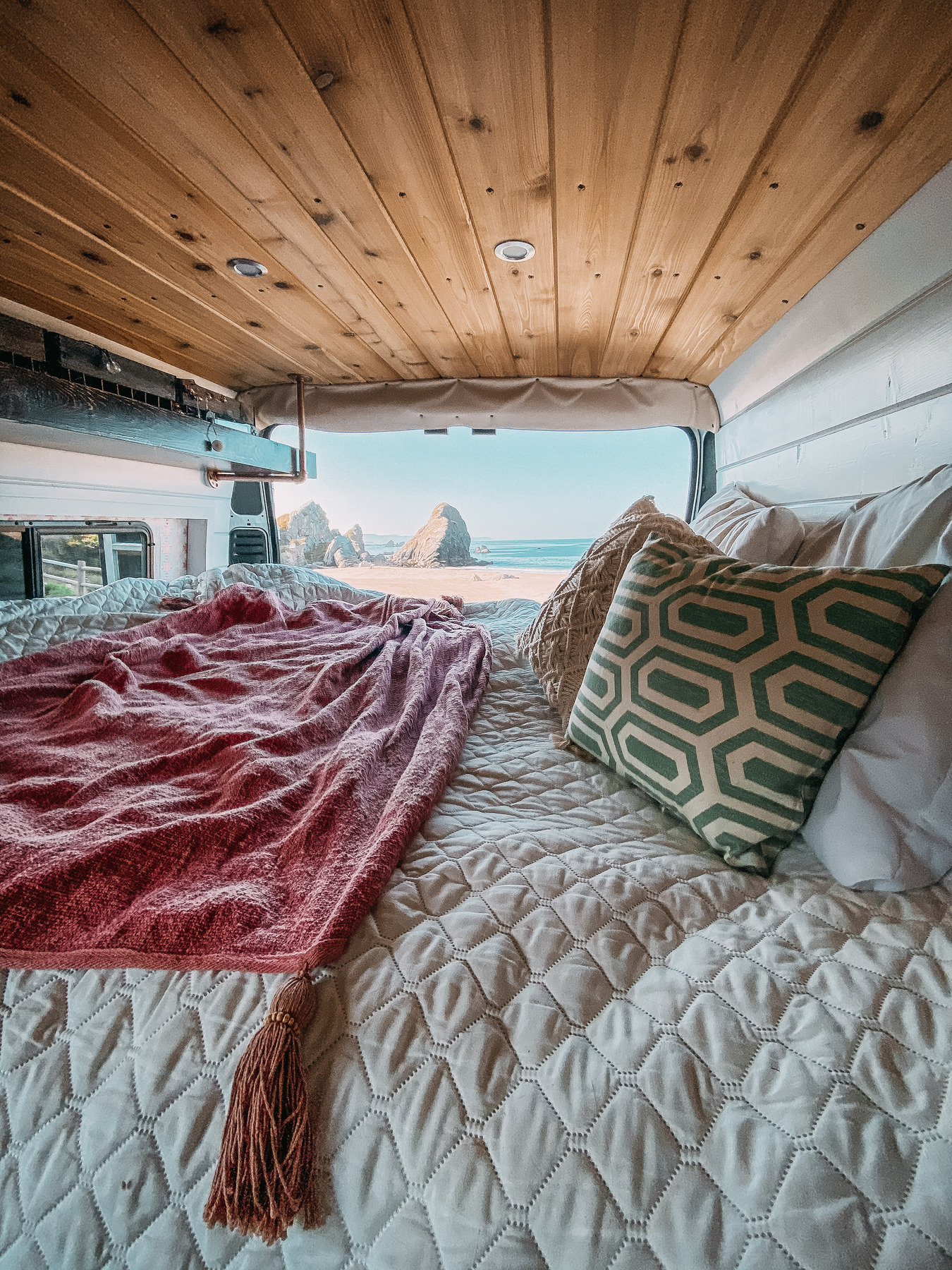 Cozy bed with pillows and a patterned blanket inside a van, wooden ceiling above, back doors open to a scenic view of sea stacks, sandy beach, and blue sky outside.