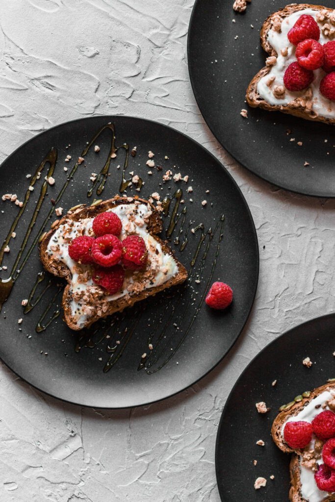 A slice of toast topped with yogurt, granola, and fresh raspberries sits on a black plate with a honey drizzle and a single raspberry on the side; two more similarly prepared plates are partially visible.