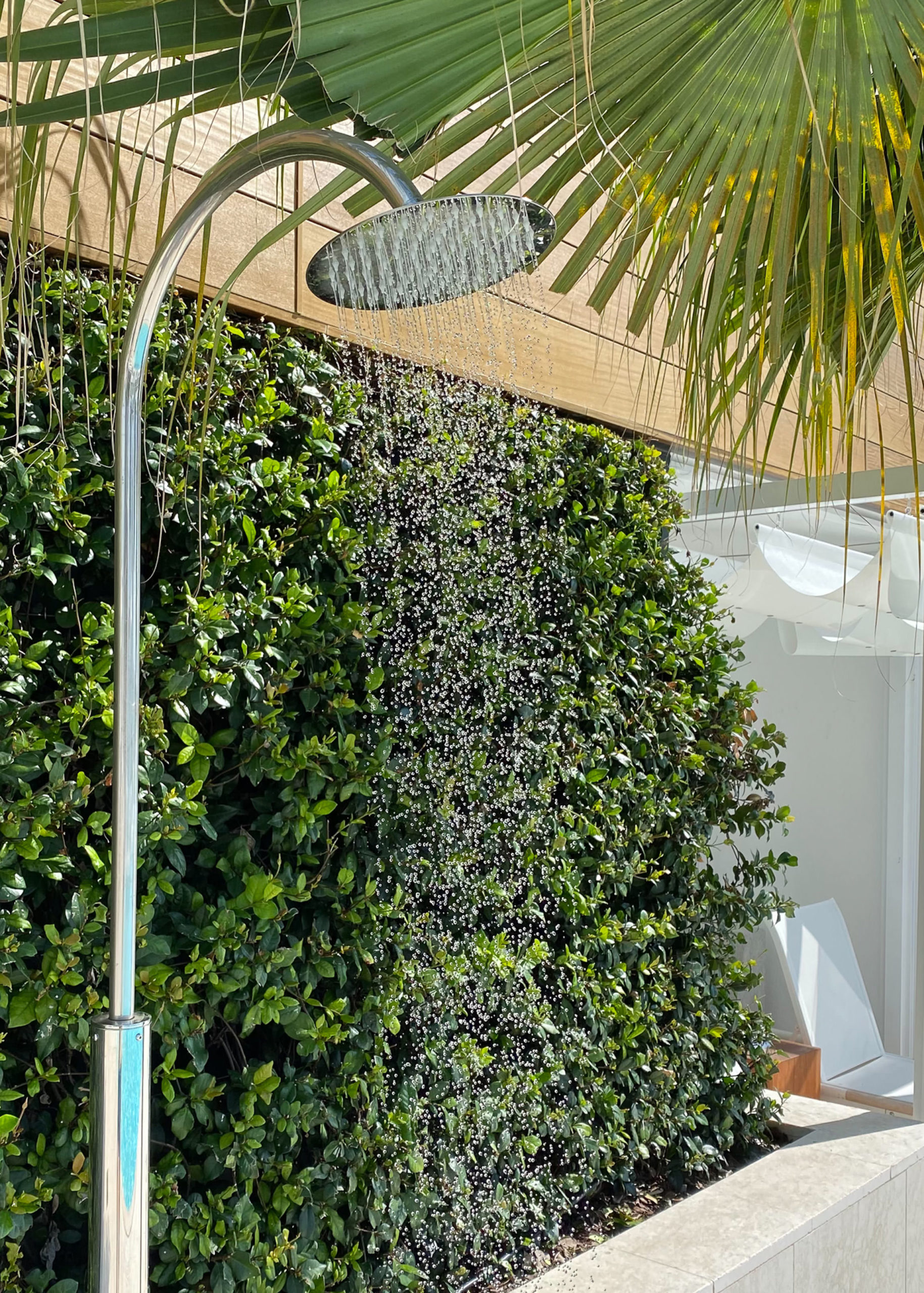 A modern outdoor shower with water flowing, set beside lush green bushes and shaded by large palm leaves under a sunlit canopy.