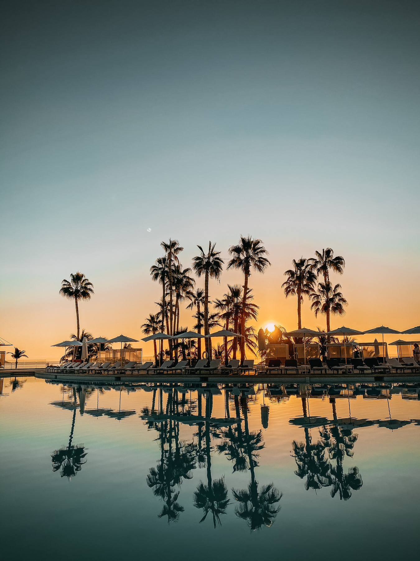 A row of palm trees and lounge chairs by a pool at sunset, with the sky glowing orange and the trees reflected in the calm water. Umbrellas and silhouettes of people are visible in the tranquil scene.