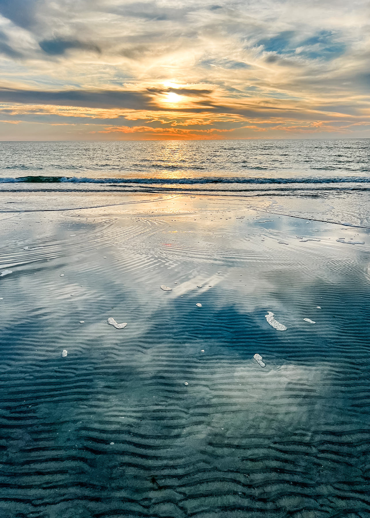 Sunset over a calm ocean, with golden light reflecting off wet sand and shallow water on the beach. Ripples in the sand and soft waves create a peaceful, serene scene under a partly cloudy sky.