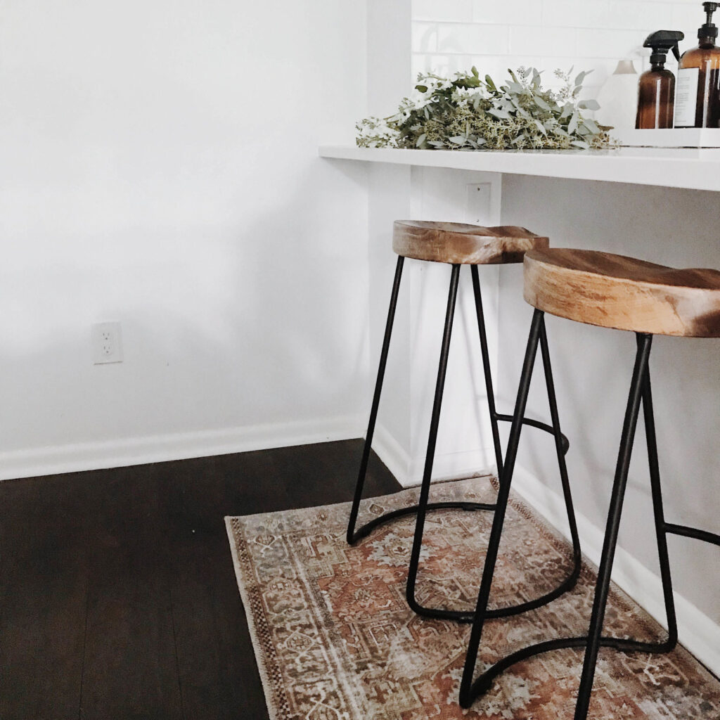Two wooden bar stools with metal legs sit at a white countertop. A vintage patterned rug lies on dark wood flooring, and greenery with brown bottles decorates the counter in a minimalist, bright space.