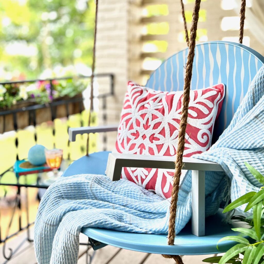 a swing made from two repurposed satellite dish is shown hanging on a balcony with a light throw and a pillow in the swing. A bit of green plant is off to one side and a flower box is on the other