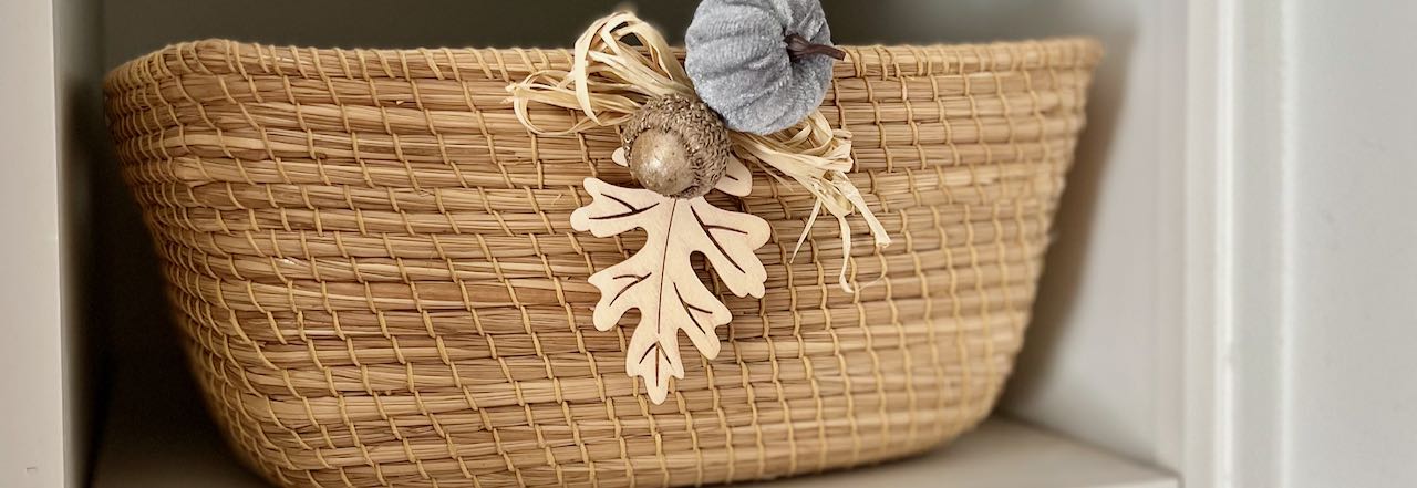 Wide Closeup of a Fall accent of wood leaf, mini velvet pumpkin, a bronzed acorn and raffia attached to tightly woven basket 