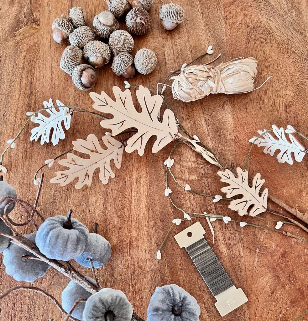 Supplies laid out on a wood table: bundle of raffia, wood leaves, velvet pumpkins, bronzed acorns, paddle of wire