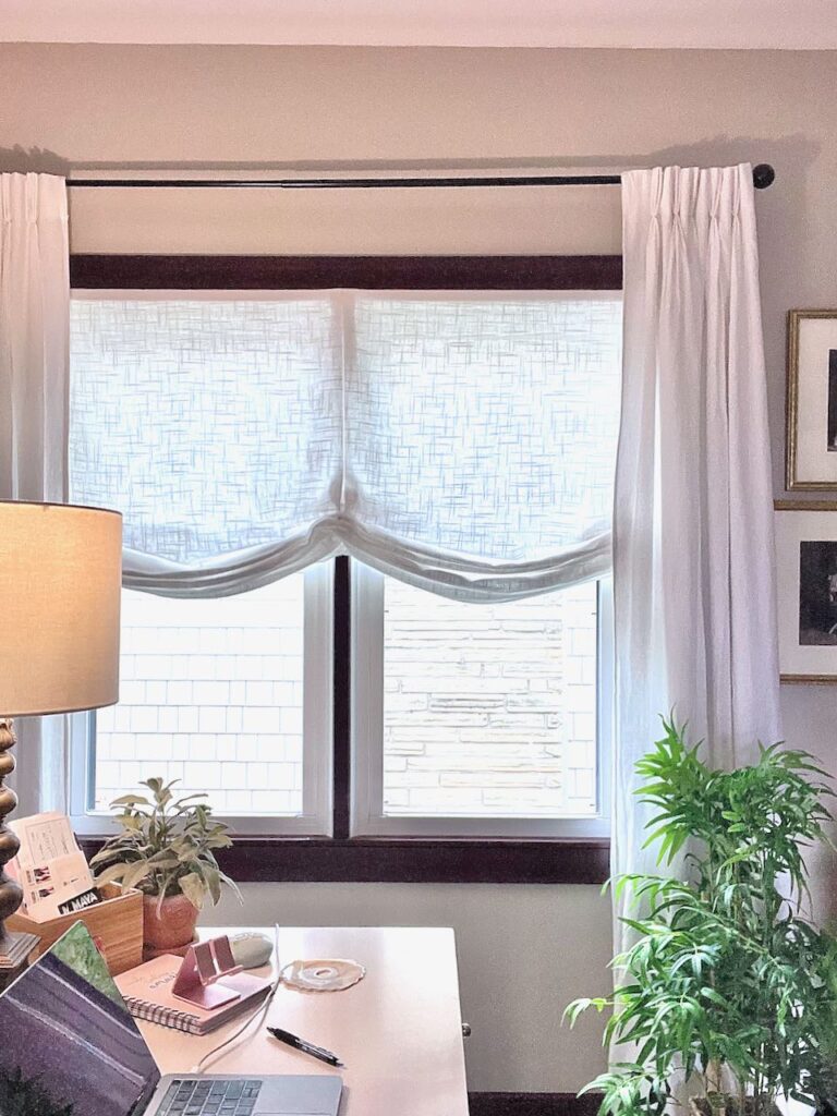 A double window with a white Roman shade and white linen drapery panels hanging on each side behind the corner of a desk