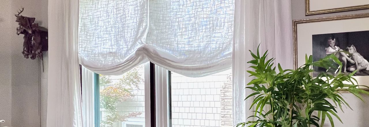 Title image closeup of a white Roman Shade in a double window with white linen drapery panels hanging on each side