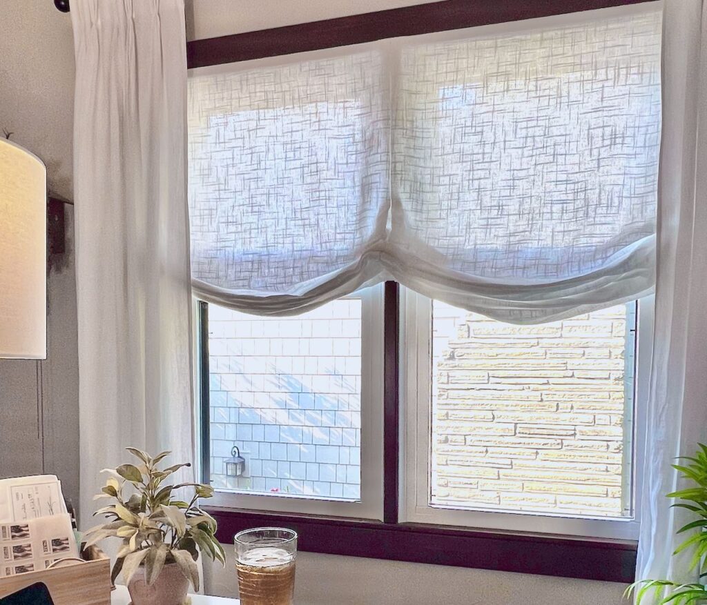A roman shade in a double window covering the upper half with linen drapery panels hanging on each side.