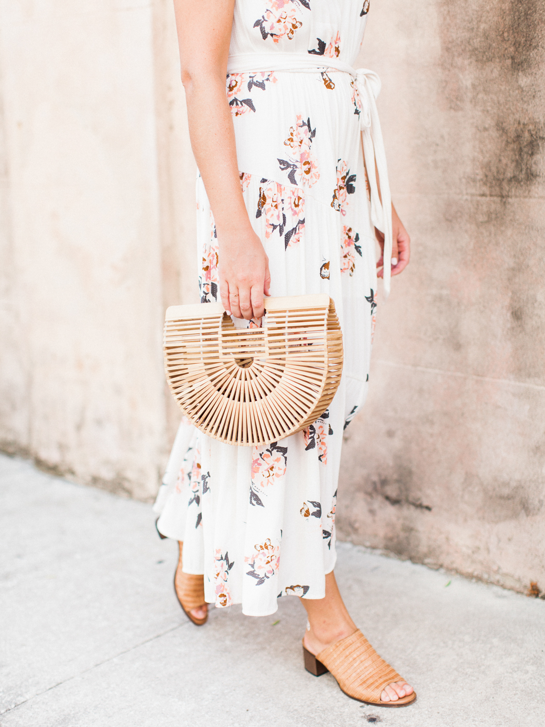 A woman in a white floral dress and tan sandals carries a semi-circular bamboo handbag while walking on a sidewalk next to a light-colored wall.