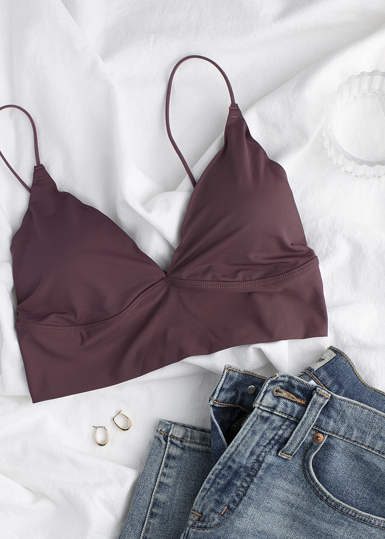 A mauve bralette with thin straps is laid on a white fabric surface next to a pair of blue jeans, two gold hoop earrings, and a clear bangle.