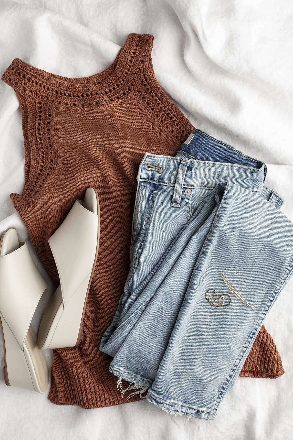 A sleeveless rust knit top, light blue jeans, beige heeled sandals, and a pair of gold hoop earrings are neatly arranged on a white fabric background.
