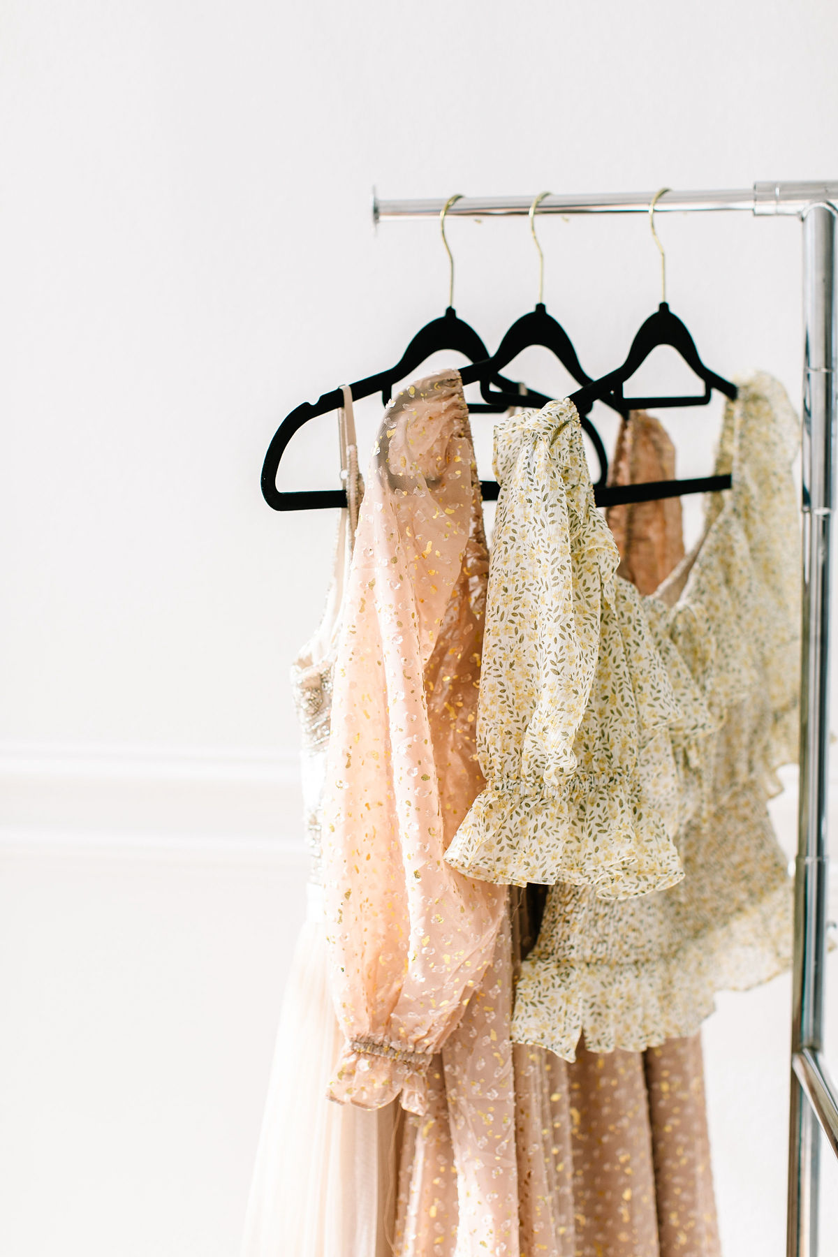 Three light, floral-patterned dresses with ruffles and puffed sleeves hanging on black hangers from a silver clothing rack against a white wall background.