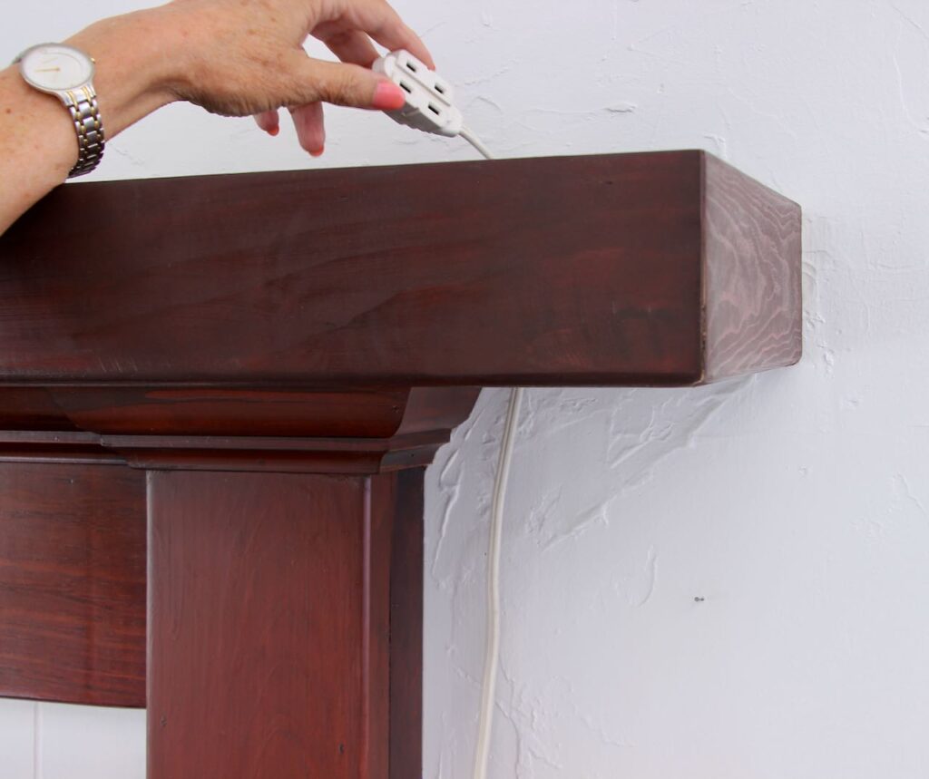 Woman's and holding an extension cord extending out between the wall and the mantel cover
