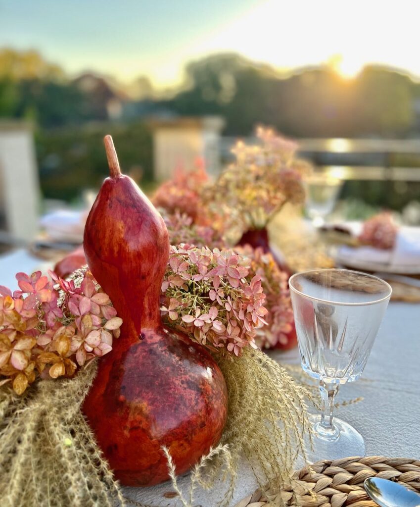 Closeup of a tall shiny dyed gourd nestled in grass heads and fall tinted hydrangea blooms with the sun rising over the lake in the background