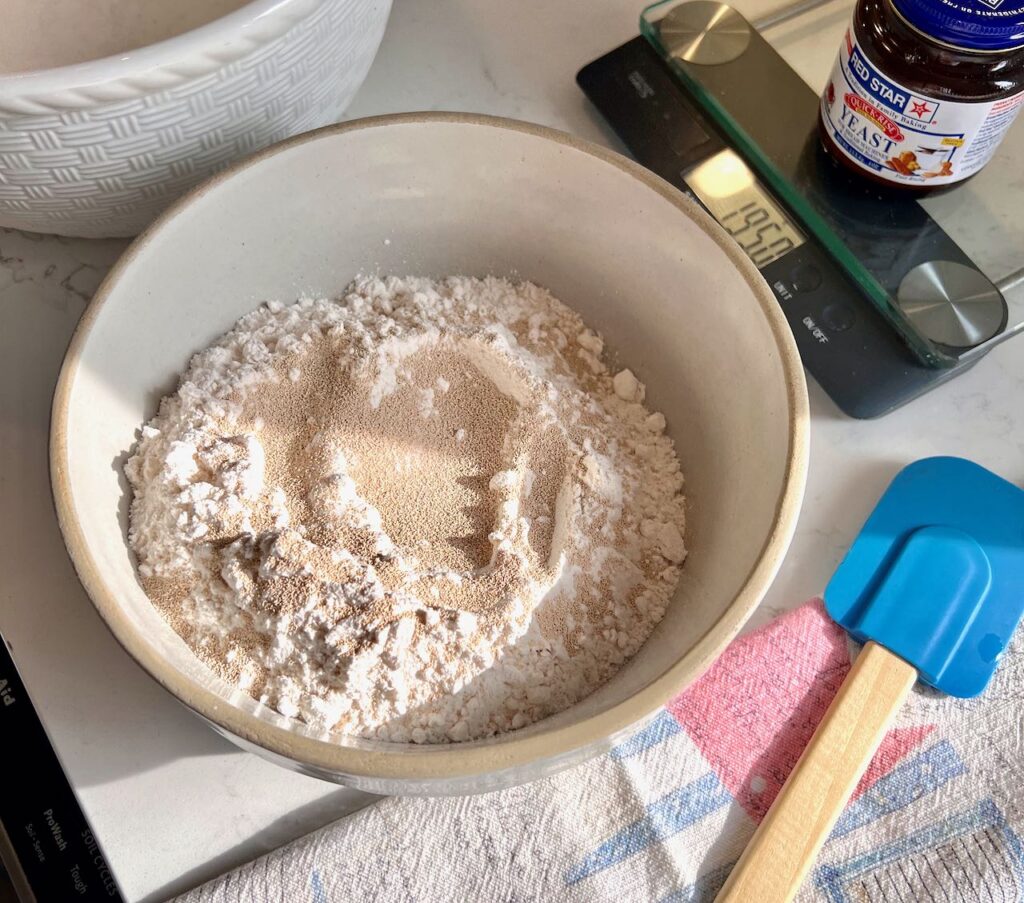 flour, salt and yeast are in a mixing bowl with a spatula beside waiting to be mixed together egther