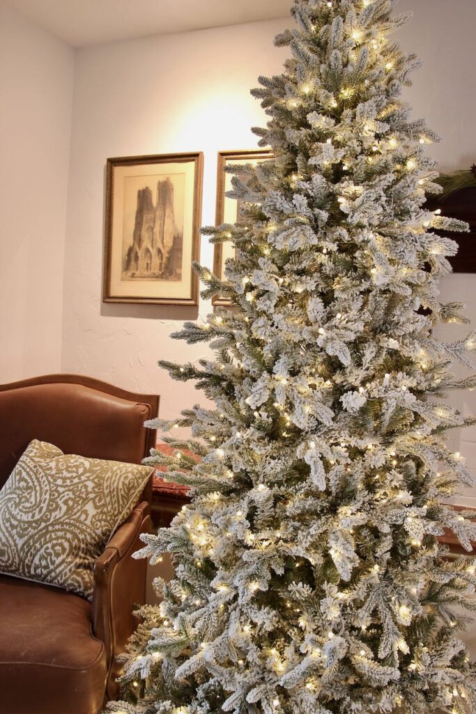 A full length image of a lit, flocked Christmas tree next to a chair