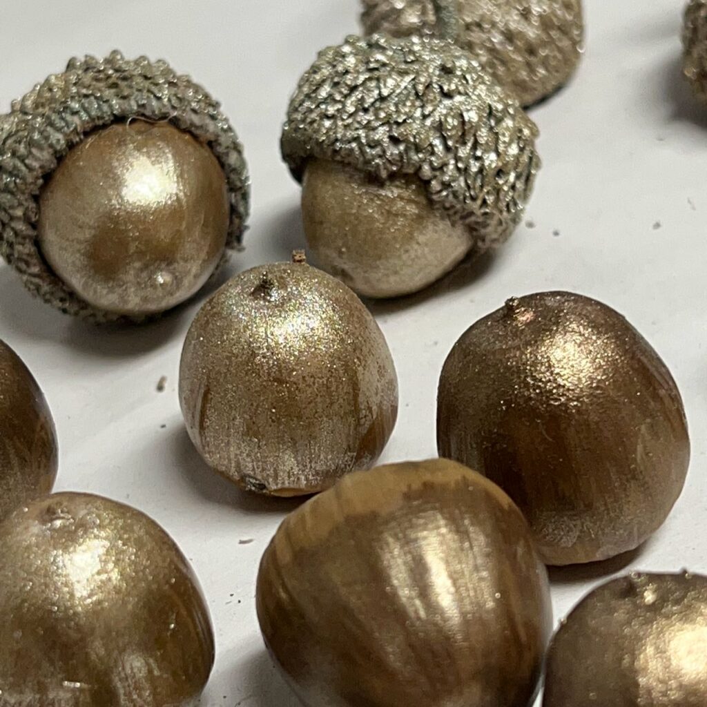 Loose acorn nuts painted metallic bronze, some with their caps painted champagne gold on a sheet of newsprint