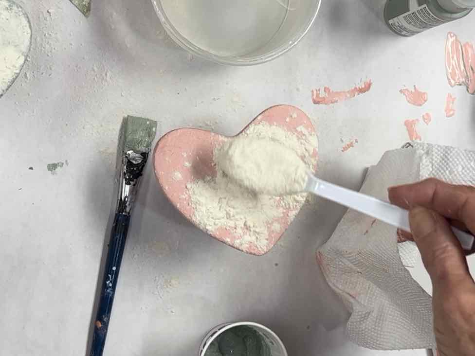 closeup of flour in a spoon being sprinkled on the wet textured paint of the clay heart