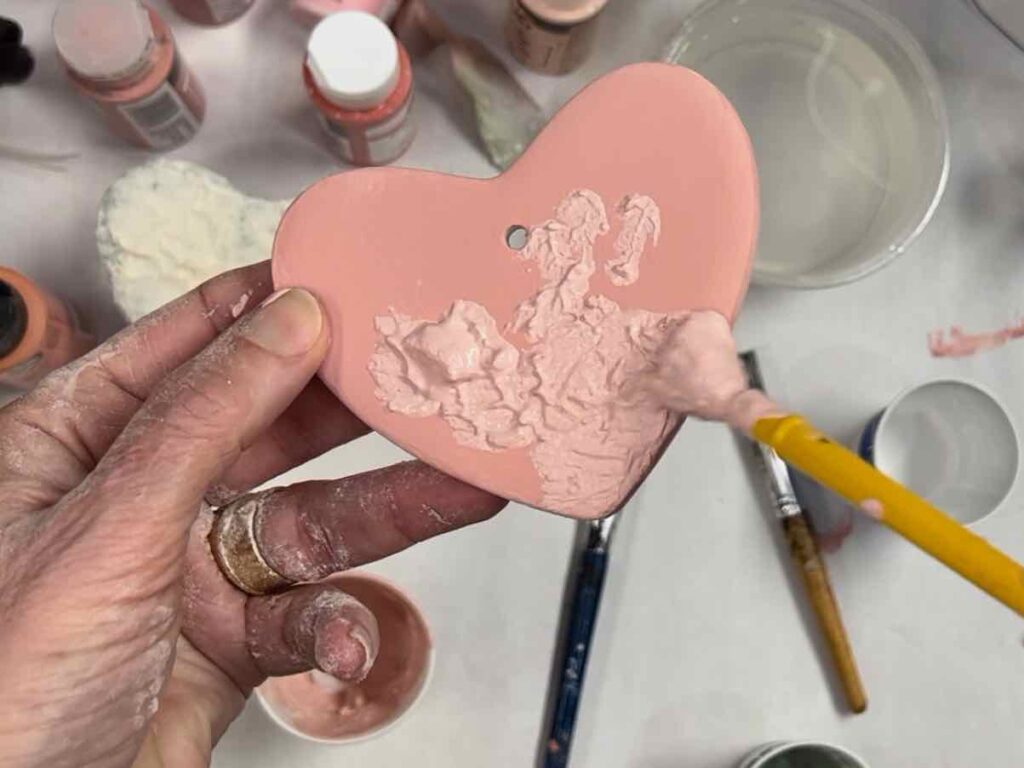 the end of a paint brush pouncing textured paint over the dry base paint on the clay heart in a woman's hand.