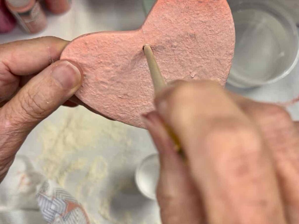 woman's hand poking the end of a thin paint brush handle into the hole of the clay heart clearing out any paint.