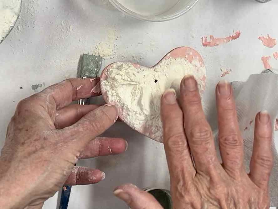 Women's fingers patting flour into the wet paint on the face of the painted clay heart