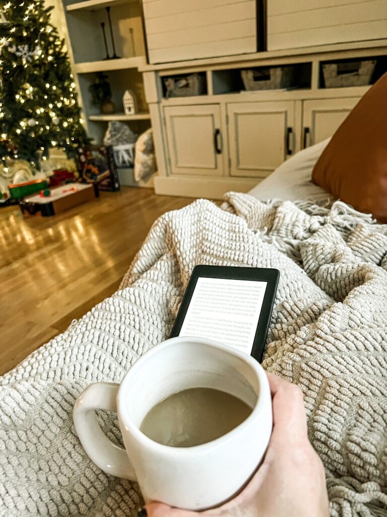 close up of a woman's hand holding a cup of coffee with a ebook on a lap throw and a lighted tree in the corner