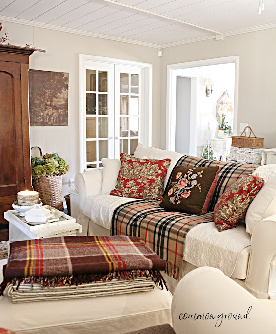 Plaid wool blanket is spread over a neutral couch with floral throw pillows and a stack of wool blankets are on a footstool