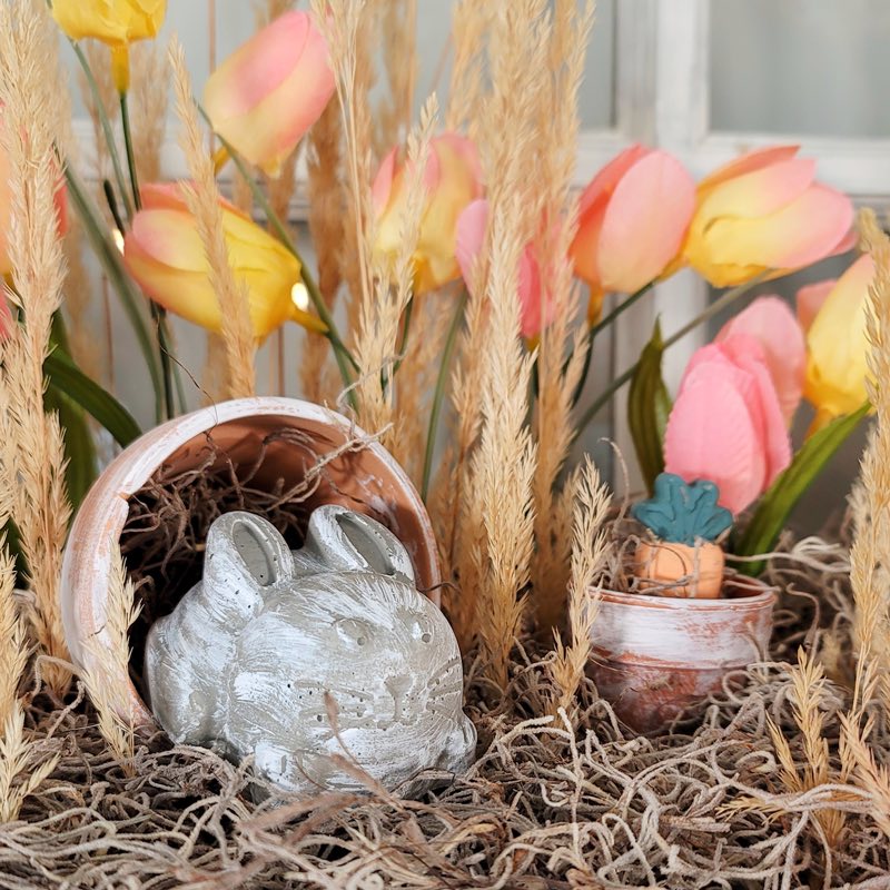 little concrete bunny is peeking out of a small flower pot in a floral arrangement of tulips and moss