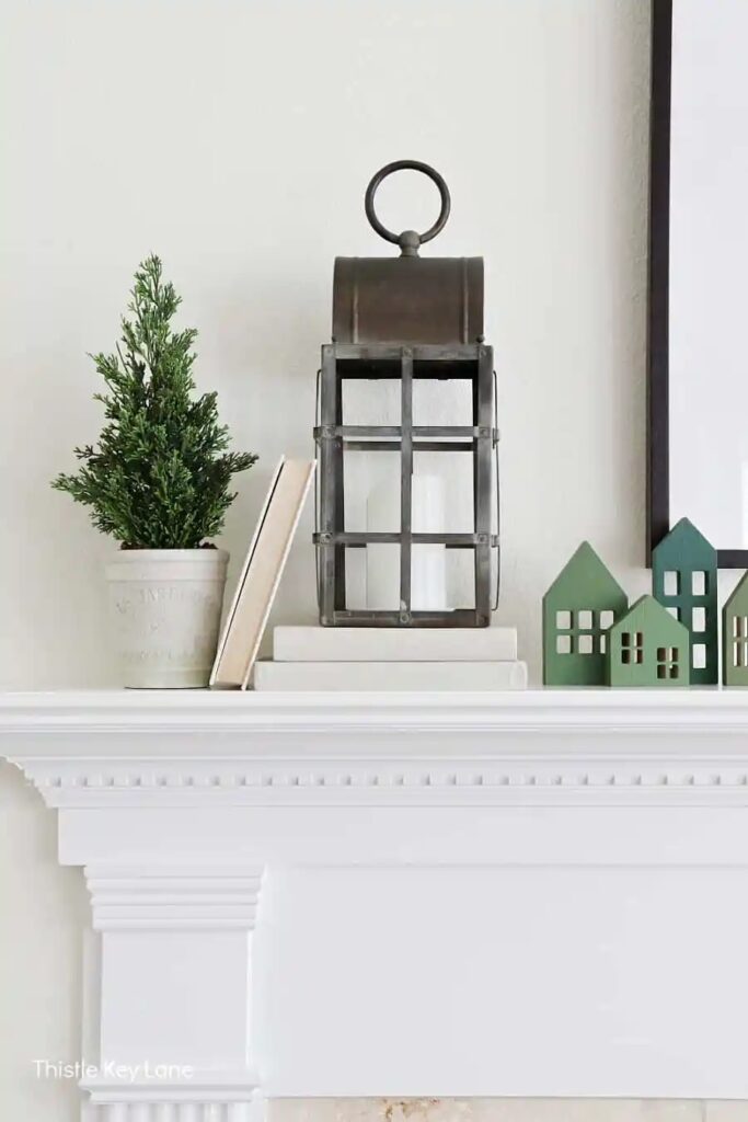 the end of a white mantel and fireplace surround with. a black lantern on some books with greenery in a vase and little green houses beside