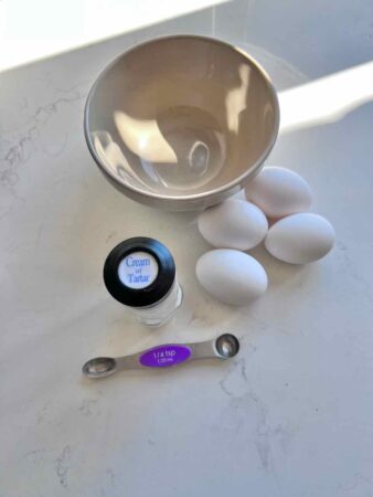 a small mixing bowl on a counter with four eggs and a bottle of Cream of Tartar and a measuring spoon