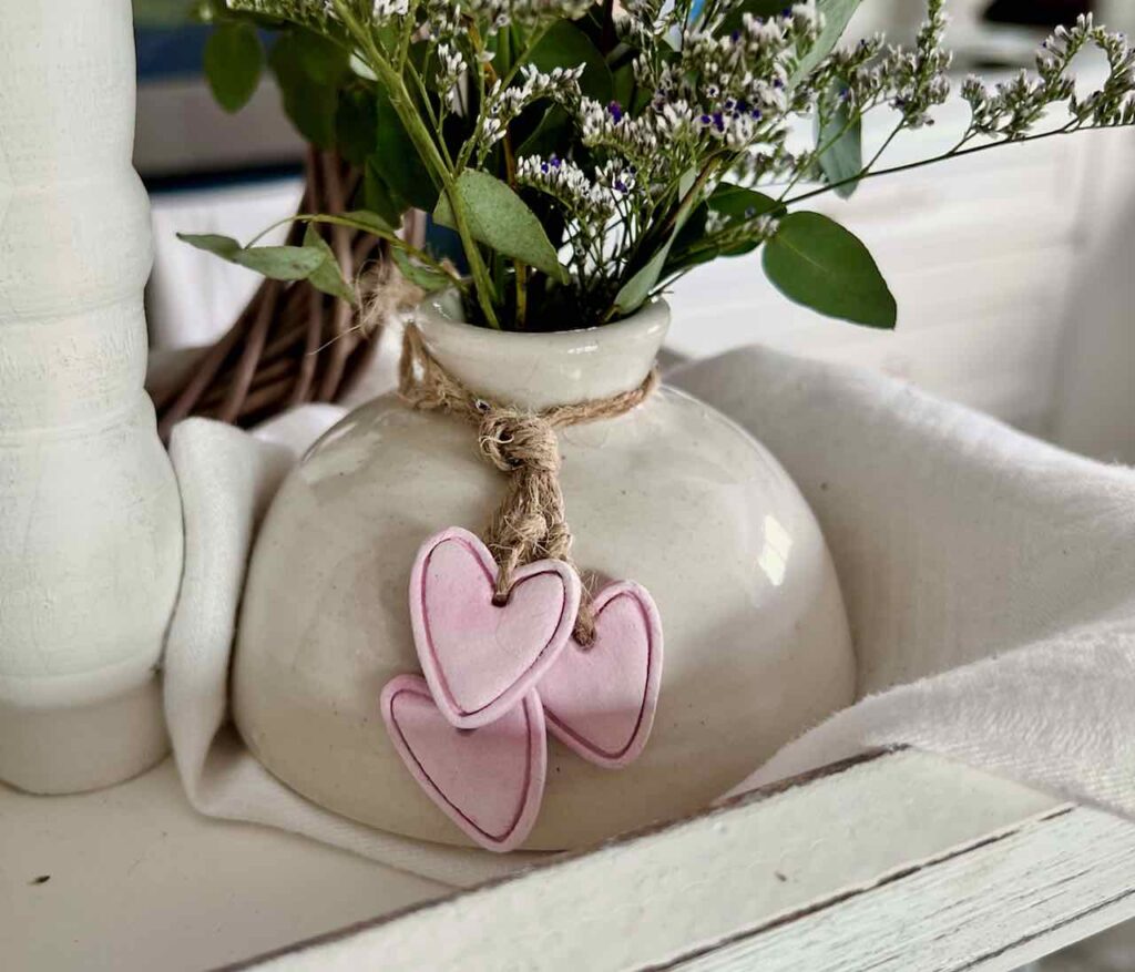 Three small clay hearts hanging from jute are tied around the neck of a ceramic flower vase