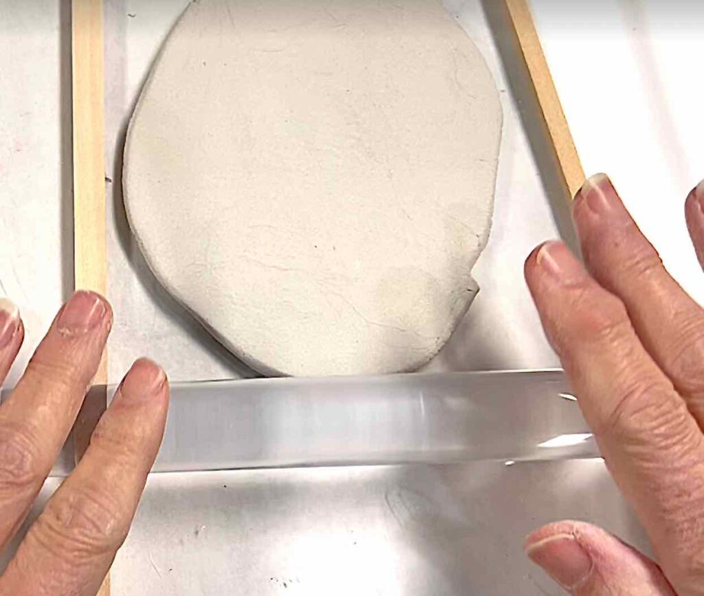 fingers shown rolling a clear acrylic rod over air dry clay between two sticks of wood