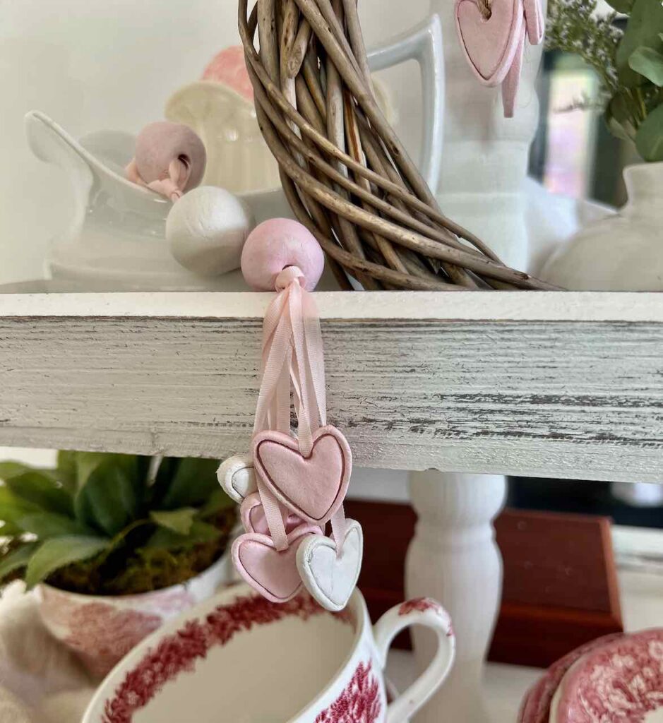 six small clay hearts are dangling from silk embroidery ribbon threaded through clay beads as a tassel hanging from a tiered tray