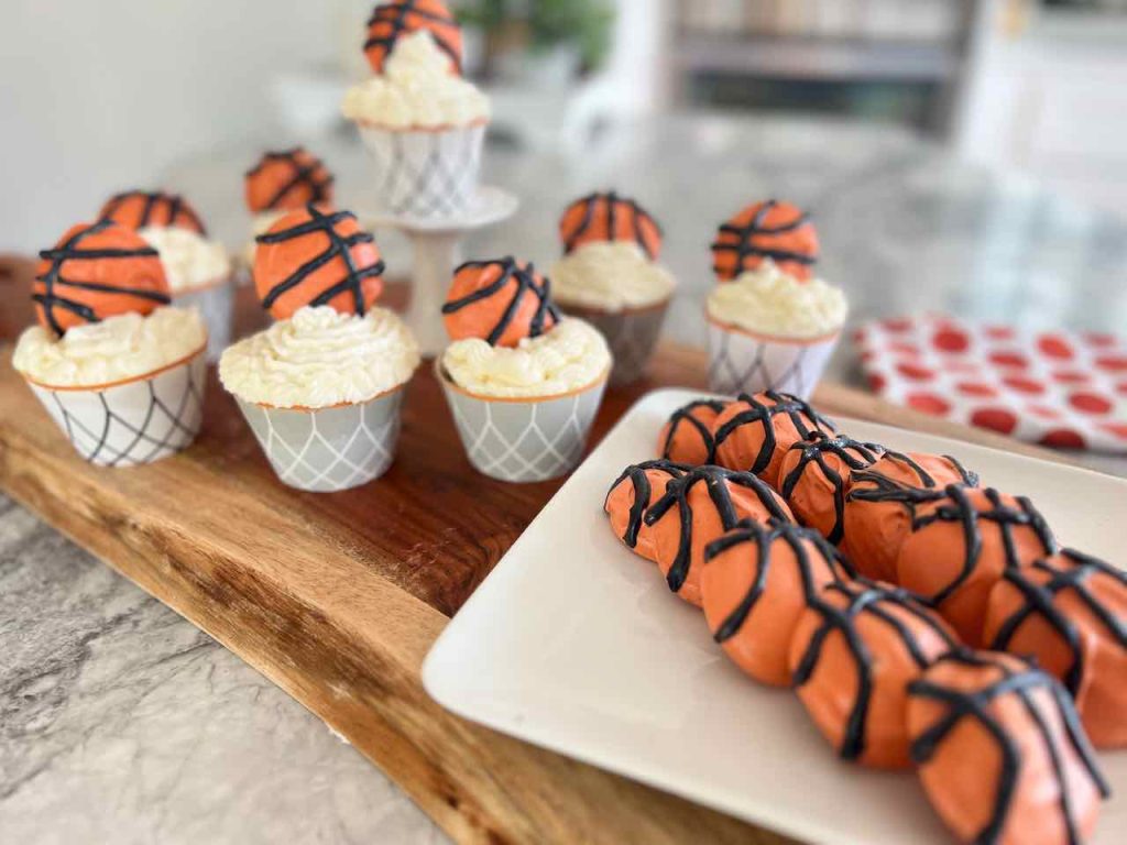 A plate of basketball cookies on a wood serving board with basketball cupcakes behind