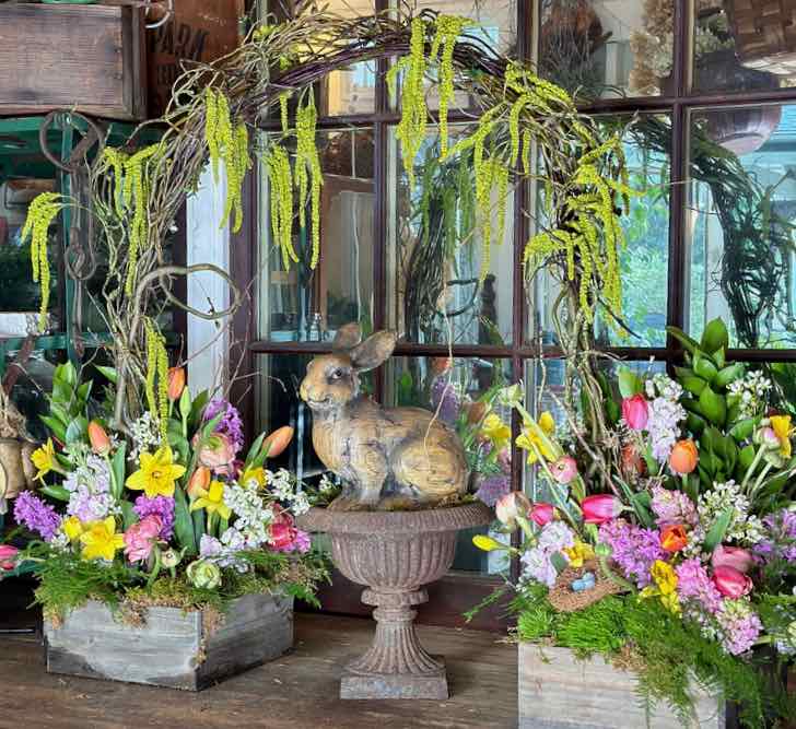 An Easter flower display of two arrangements flanking a bunny statue under and arch in front of a vintage mirror