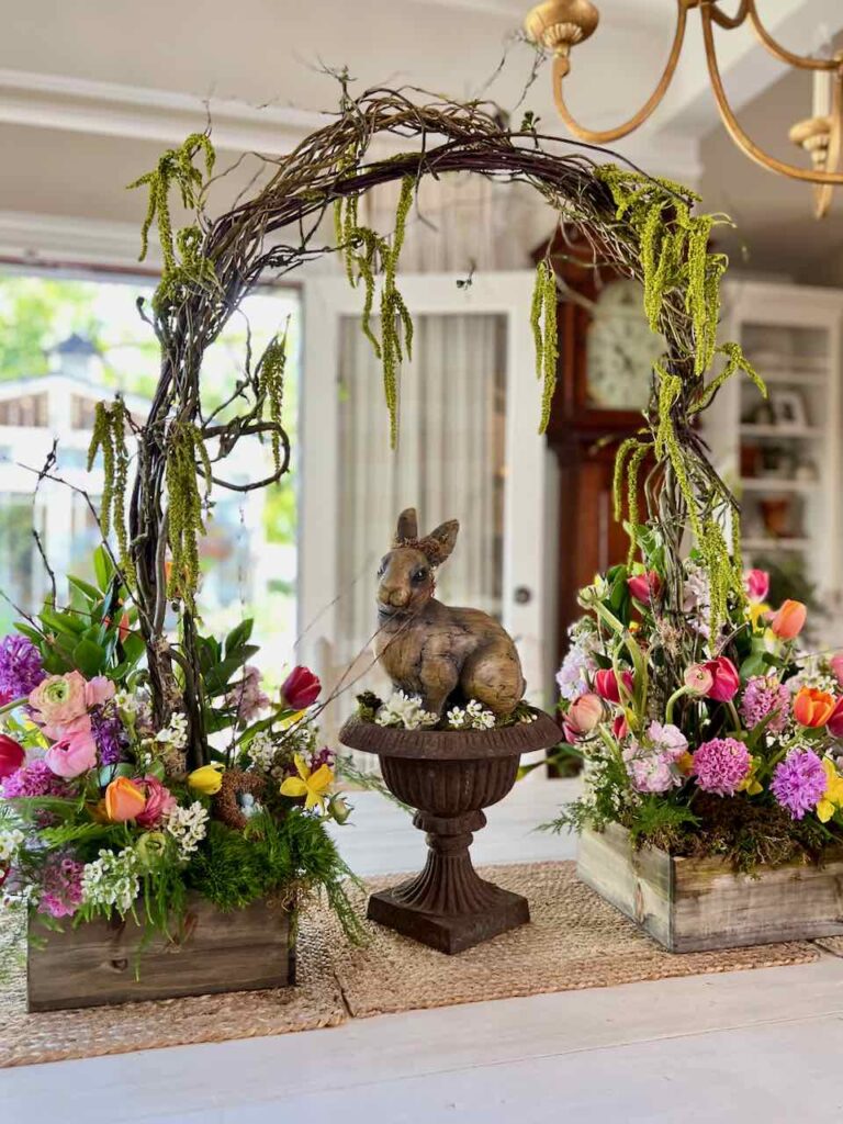 An Easter flower display of two arrangements flanking a bunny statue under and arch