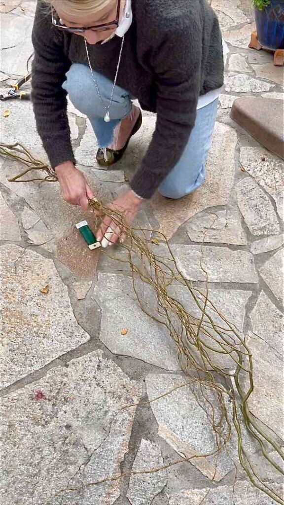 Woman kneeling on patio wrapping florists wire around long bundle of willow branches