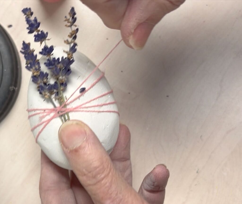 Hand holding flowers on egg while other hand wraps twine around the egg
