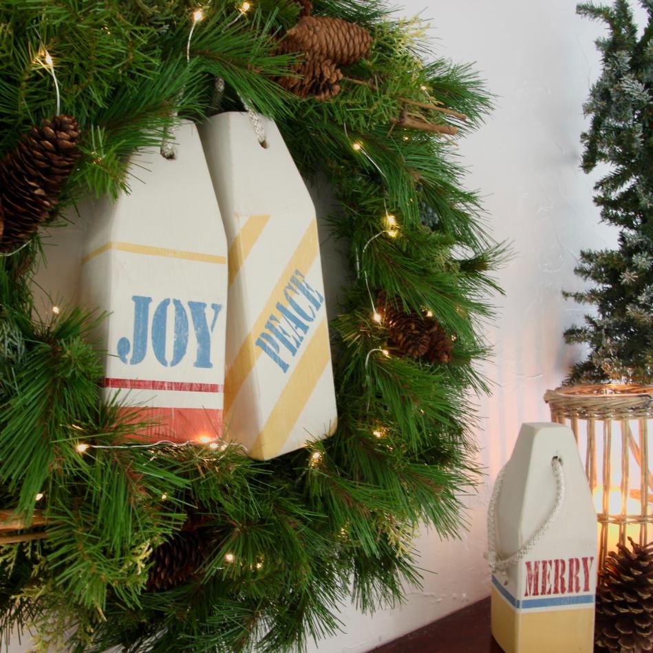 A Christmas wreath with pinecones and two wooden buoys labeled "JOY" and "PEACE." Nearby, another buoy with "MERRY" sits next to a small decorated tree.